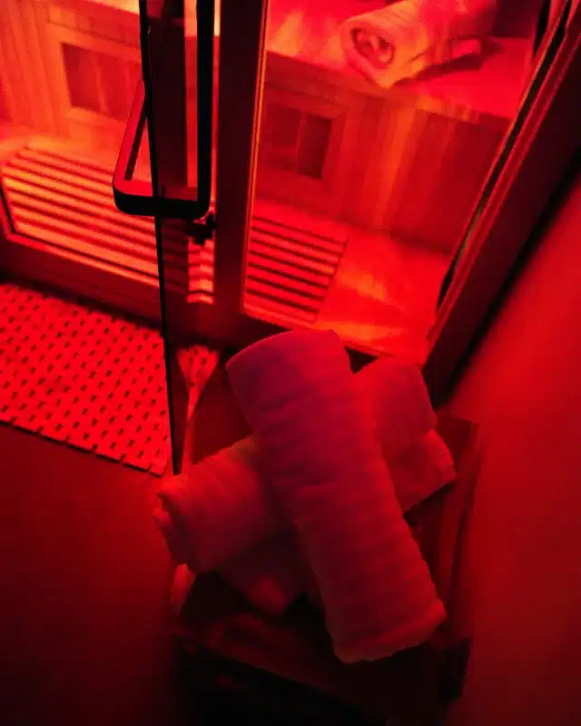 Rolled white towels outside glowing red infrared sauna cabin with wooden interior at fitness studio