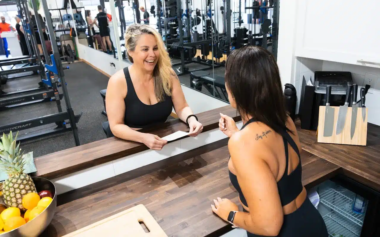 Smiling blonde trainer hands document to dark-haired client at juice bar counter in fitness studio
